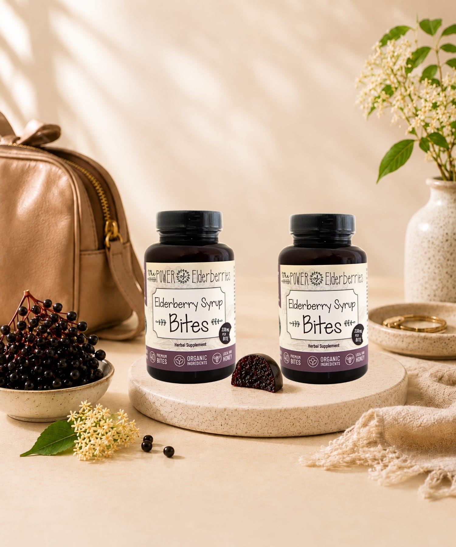 Two bottles of The POWER of Elderberries® Elderberry Syrup Bites displayed with elderberries, ginger root, and local raw honey in a light luxury wellness setting.