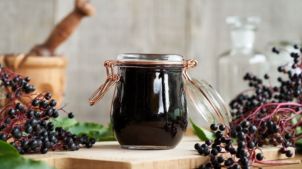 Glass jar of elderberry syrup surrounded by fresh elderberries and herbs on a wooden surface