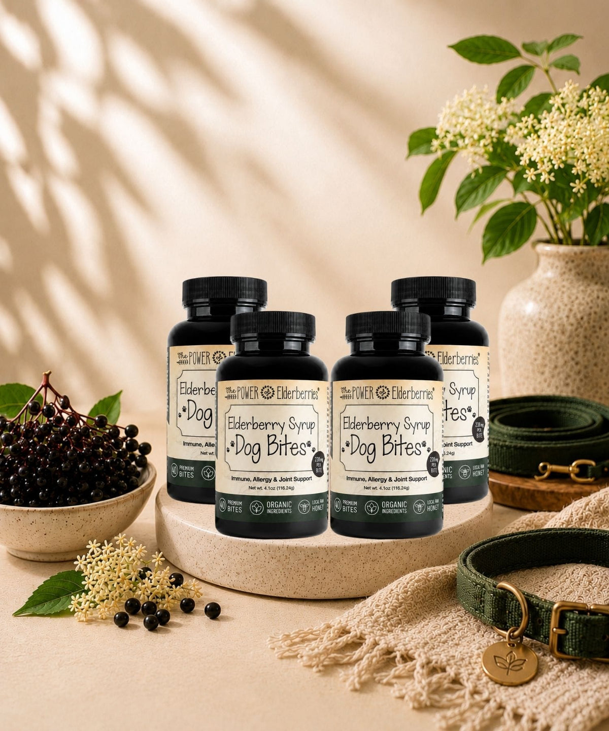 Four bottles of The POWER of Elderberries' Elderberry Dog Bites arranged on a round stone tray, styled with a green collar, leash, elderberries, elderflowers, and a textured blanket in a warm indoor setting.