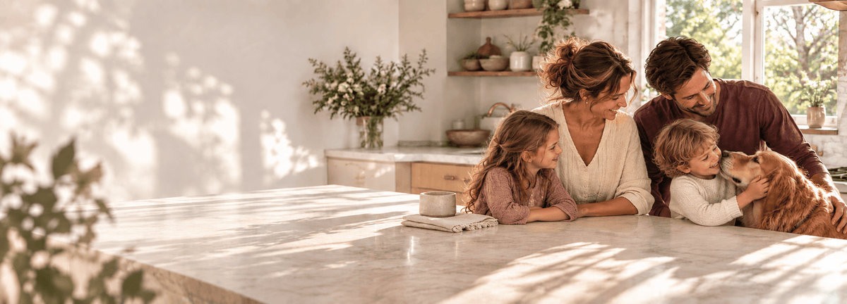A family of four and their dog relax at a white marble kitchen island in a bright, modern kitchen with warm natural light.